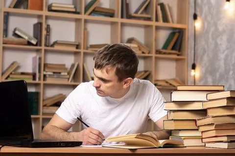 Young student writes an essay at the table with books and looks into a laptop. Stock Photos
