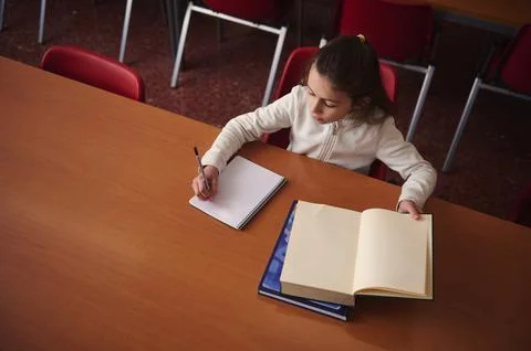Young Student Writes Notes At Table With Open Book In Classroom Library Stock Photos