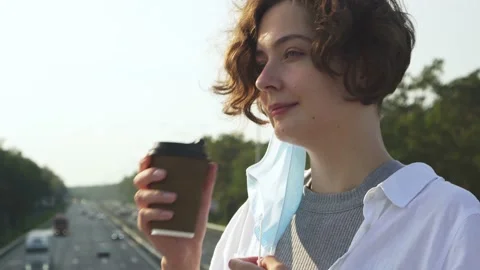 Young stylish brunette in mask drinks coffee on footbridge. Beautiful young Stock Footage 161040642