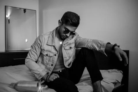 A young stylish guy in a denim jacket on the bed with a bottle of alcohol. bl Stock Photos