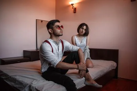 A young stylish guy in glasses and a young girl together sitting on the bed.  Stock Photos