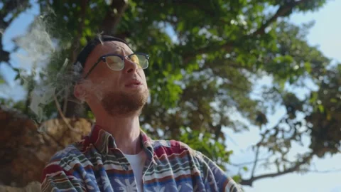 Young stylish guy relaxes smoking marijuana outdoors under a tree in summer Stock Footage 237042736