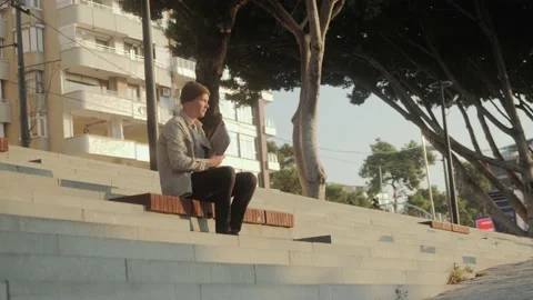 A young stylishly dressed man is sitting on the steps during sunrise Stock Footage 221582938