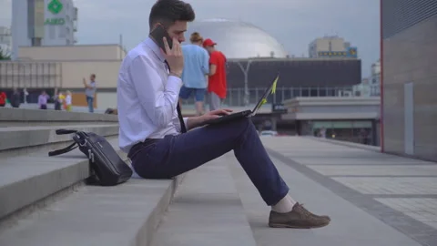 Young successful guy student working on the street on laptop. A man in a white Stock Footage 91419391