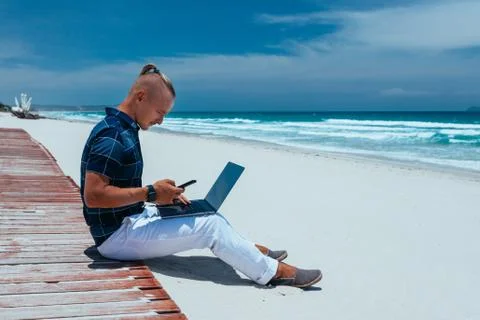 Young successful guy working using a laptop, sitting on the seashore Stock Photos
