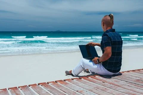 Young successful guy working using a laptop, sitting on the seashore on a san Stock Photos