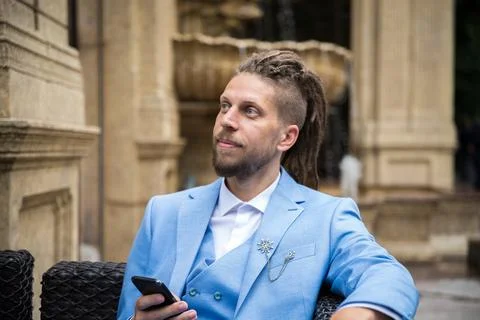 A young successful man in a blue suit with a smartphone in his hands sits in a Stock Photos