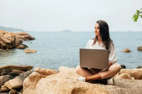 Young successful, smiling girl working using a laptop, sitting on a rocky sea Stock Photos