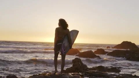 A young surfer with a surfboard in her hand, lying on a rock in the ocean Stock Footage 243180841