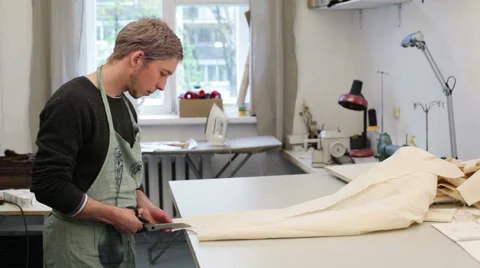 A young tailor man in an apron cuts a piece of fabric large scissors Stock Footage 62672936
