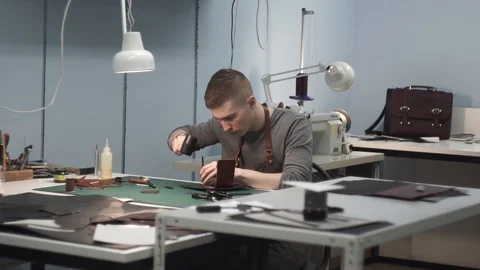 A young tanner in an apron sits at a table in the workshop. He punches holes in Stock Footage 104925296