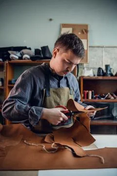 A young tanner in his workshop at the table cuts out leather elements of shoes 스톡 사진