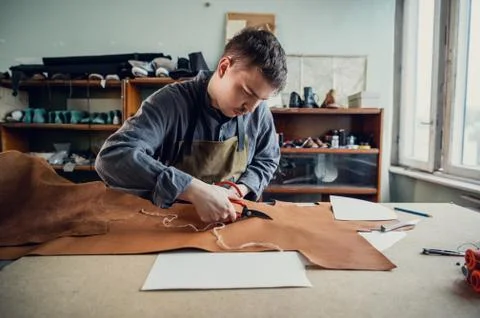 A young tanner in his workshop at the table cuts out leather elements of shoes 스톡 사진