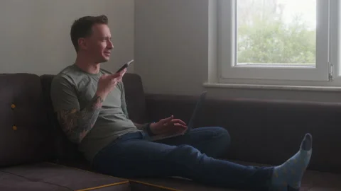 Young tattooed guy sits on sofa with computer on his knees and dictates voice Stock Footage 233131717