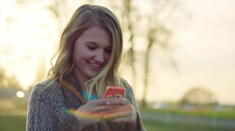 Young teenage girl texting outdoors on phone at sunset, smiling