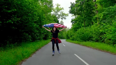 Young teenage woman with US flag is roller skating through woods and holds the Stock Footage