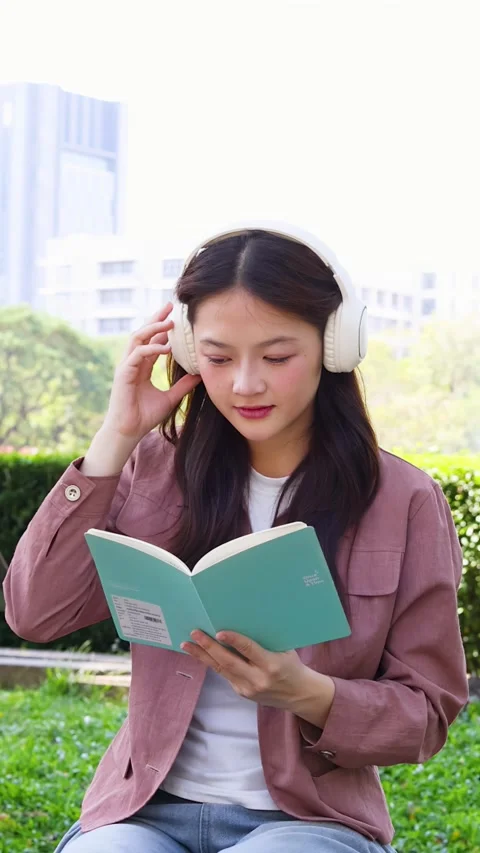 Young Thai University Student Reading a Book with Headphones Outdoors Stock Footage 328697683
