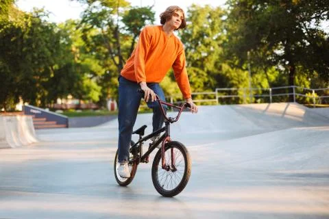 Young thoughtful guy in orange pullover and jeans dreamily riding bicycle at Stock Photos