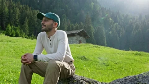 Young thoughtful man sitting on a stone in a green meadow. He is relaxing and Stock Footage 340997331
