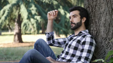 Young thoughtful man sitting under a tree at park looking at camera Stock Footage 136517588