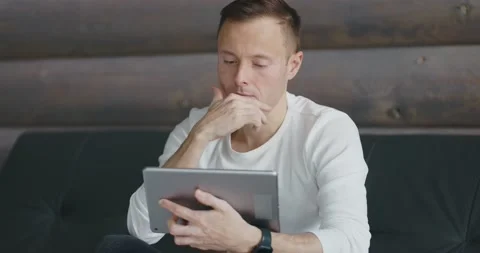 Young thoughtful man uses tablet while sitting on the couch. Front view Stock Footage 197816395