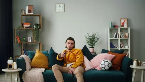 Young tired man is sitting on a sofa and looking for entertainment programs on Stock Footage 156823290