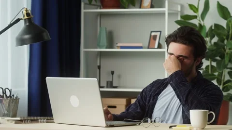 Young tired man taking off circle glasses and typing on keyboard of laptop Stock Footage 157235393