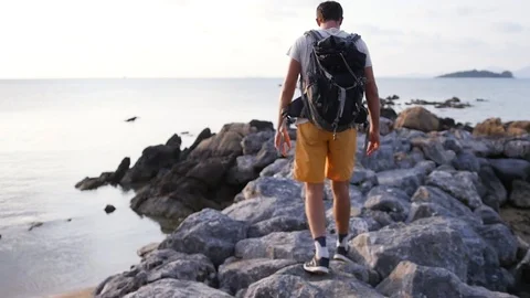 A young tourist with a backpack behind him goes along the shore of rocks against Stock Footage 93707146