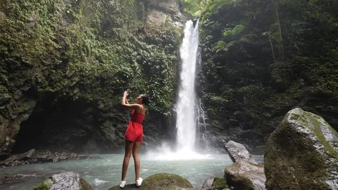 A young tourist standing at a waterfall, capturing photos using a smartphone. Stock Footage 126088199