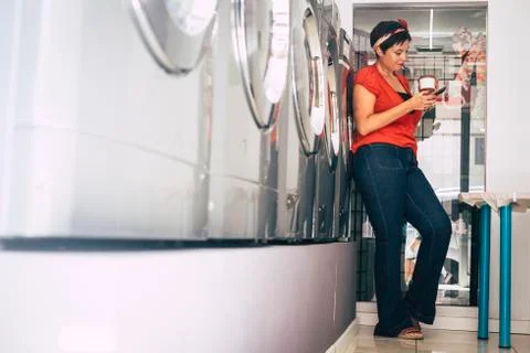 Young trendy fashion brunette use phone and wait in a laundry mat service sto Stock Photos