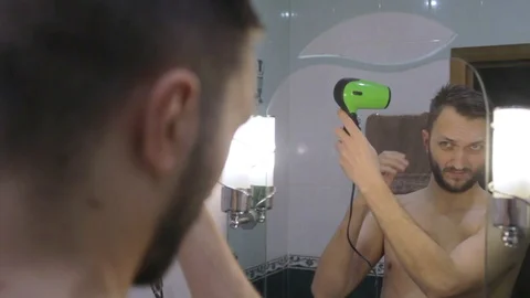 Young undressed man looking at mirror  drying hair with hair dryer Видео 104022248