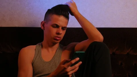 Young upset man sitting on the sofa and reading news on his smartphone at home Видео 106029147