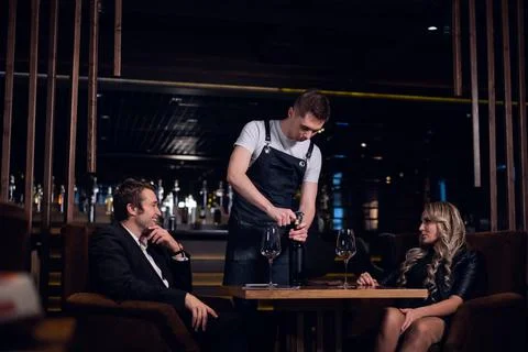 A young waiter guy opens a bottle of wine at the table in a restaurant Stock Photos