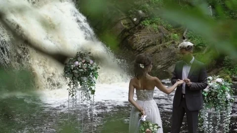 Young wedding couple exchange rings at ceremony near waterfall Stock Footage