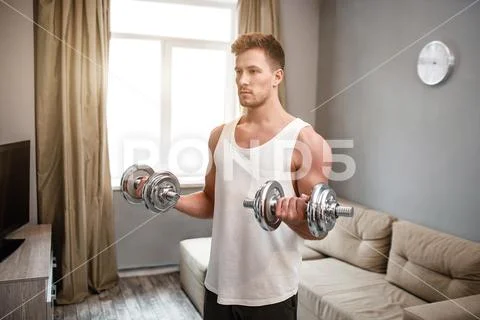 Young well-built man go in for sports in apartment. Guy doing biceps dumbbell Stock Photos
