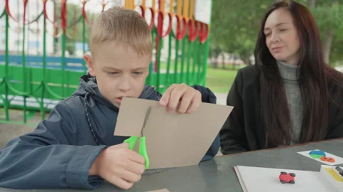 Young white boy cutting cardboard outdoors, mother watching, scissors and paint Stock Footage 345166926