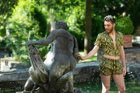 A young white guy wearing a python set of shorts and a shirt touching a sculp Photos