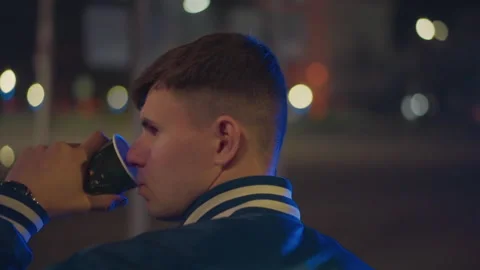 Young white man at empty street holding cup in varsity jacket profile lit by Stock Footage 329458717