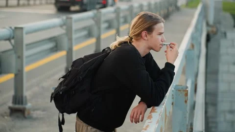 Young white man leaning bridge railing, backpacked traveler smoking cigarette Stock Footage 332365592