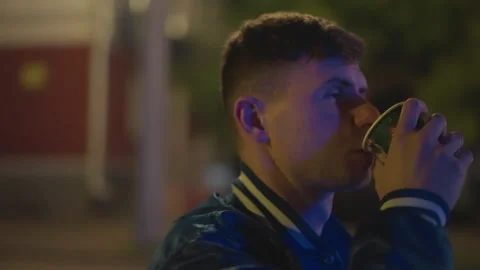Young white man standing on sidewalk sipping takeaway drink wearing varsity Stock Footage 329457227