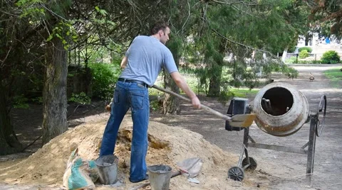 Young white worker pours sand into a rotating concrete mixer with shovel Stock Footage 64600046