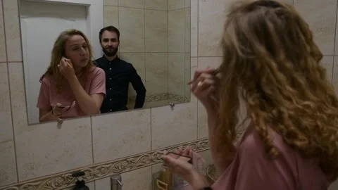 Young woman applying makeup while her husband stands behind her smiling 库存影片 330792848
