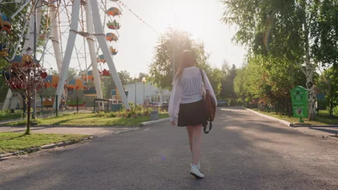 Young woman with backpack walks through sunlit amusement park path wearing Stock Footage 345844298
