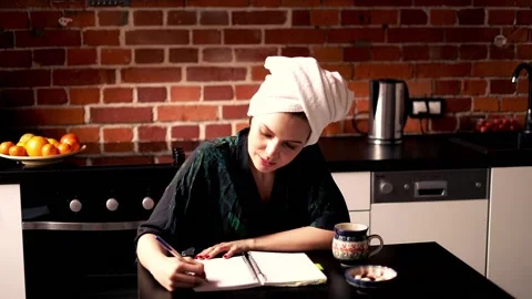 Young woman in bathrobe doing notes while sitting in the kitchen Stock Footage 90048275