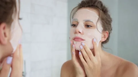 Young woman in a bathroom puts a sheet mask on her face Stock Footage