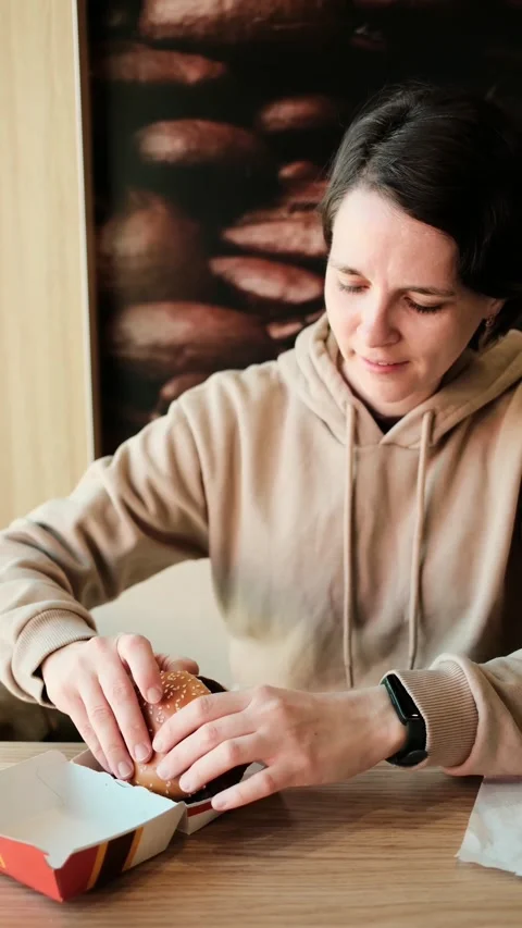 Young woman biting a cheeseburger while sitting at a table in a fast food Stock Footage 341968407