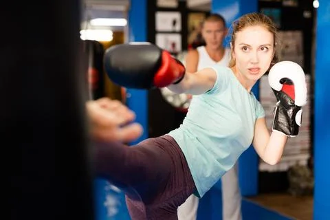 Young woman in boxing gloves practicing heel kick on punching bag in gym Stock Photos