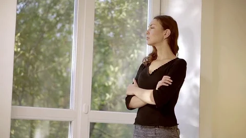 A young woman with brown hair looks out the window and touches her shoulders, ma Stock Footage