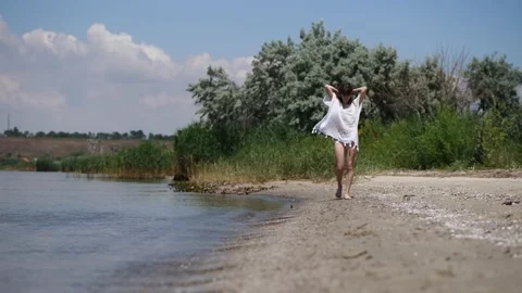 A young woman with brown hair in a white sundress is walking along the beach Stock Footage