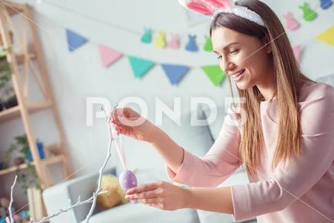 Young woman in bunny ears at home easter celebration concept hanging egg on tree Stock Photos